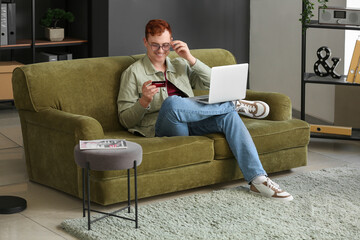 Poster - Handsome young man with credit card and modern laptop sitting on sofa while shopping online