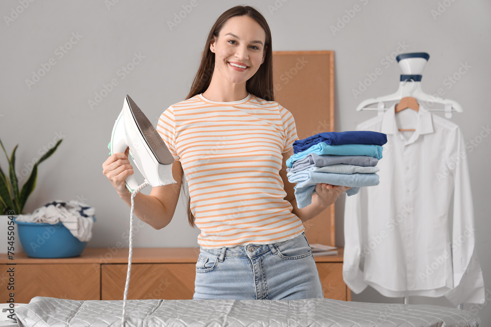 Pretty young woman with iron and stack of clean clothes at home