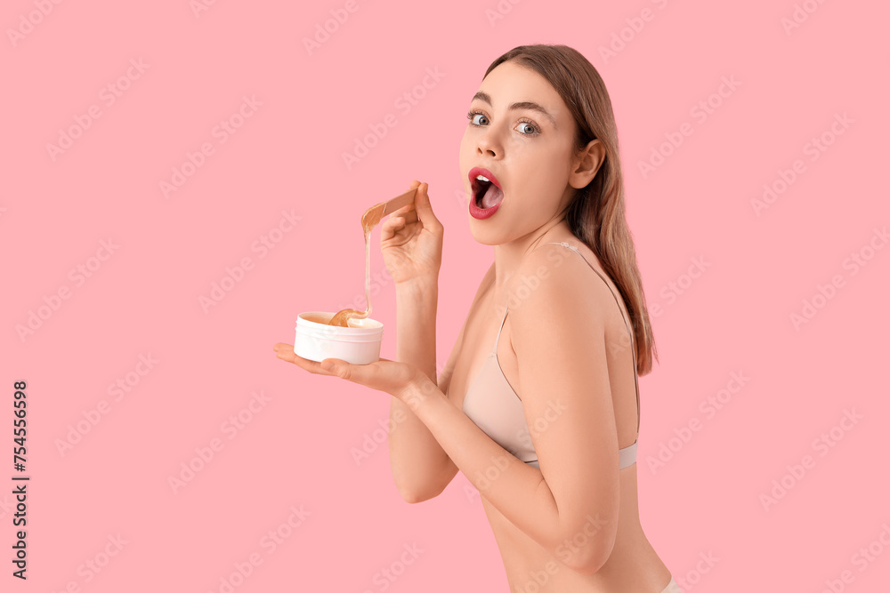 Beautiful young shocked woman holding container with sugaring paste and spatula on pink background
