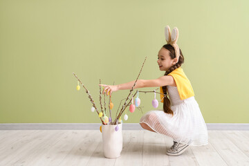 Poster - Cute little girl in bunny ears decorating willow branches with Easter eggs on green background