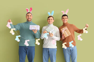 Wall Mural - Happy young men in bunny ears with gift boxes and rabbits garland on green background. Easter celebration