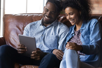 African couple watch video on digital tablet laughing have fun sit on sofa at home. Spouses spend weekend using wireless tech, virtual services, new application, buy goods, e-services online concept