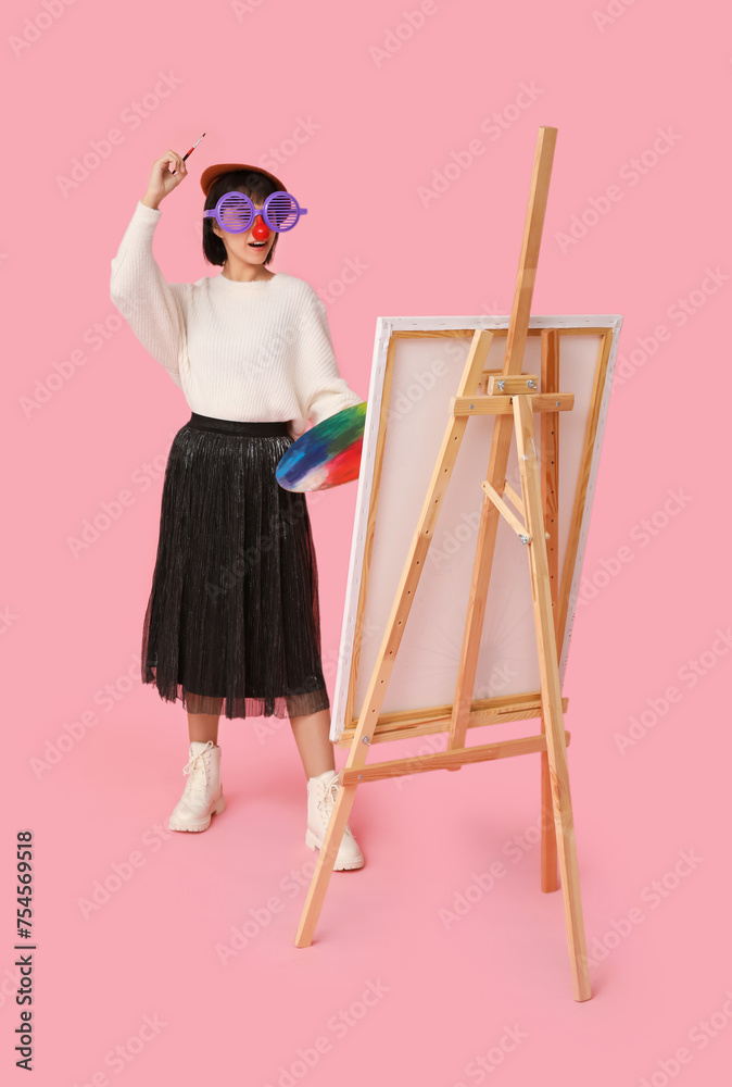 Funny young female artist with easel on pink background. Fool's day