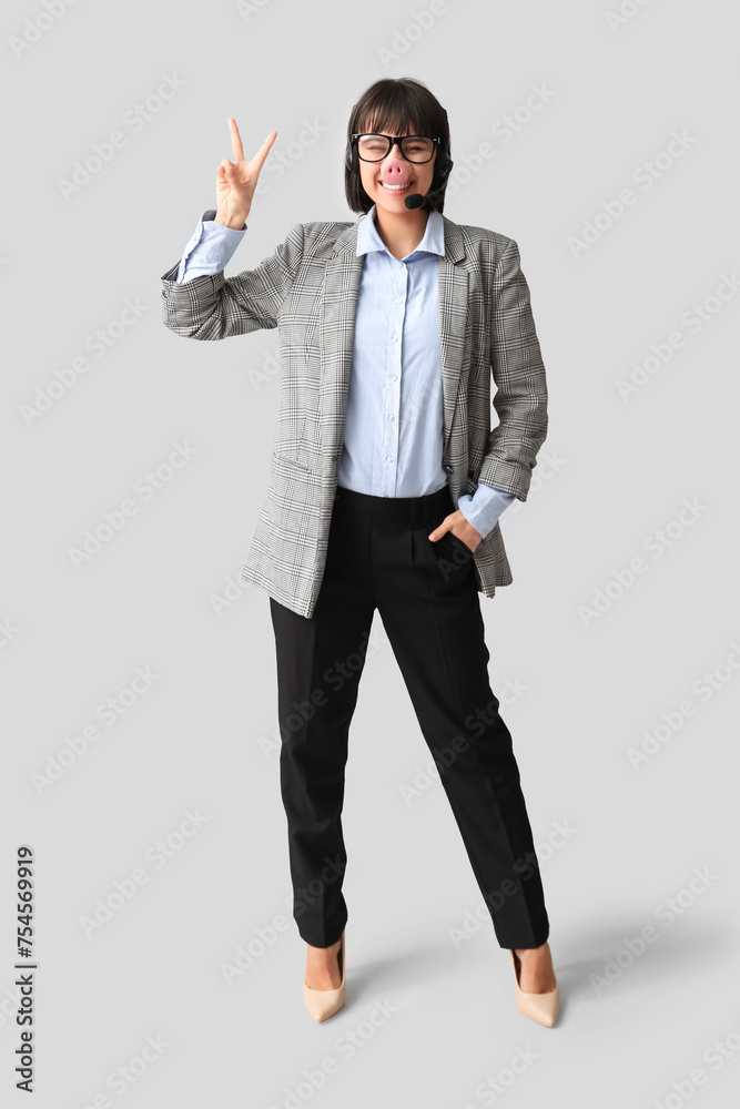 Funny young female technical support worker with headset and pig nose showing victory gesture on grey background. Fool's day