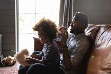 African single dad take care of little cute 6s daughter, daddy helping with hairstyle holding comb brushing her natural curly hair, loving family sit on couch do morning routine. Fatherhood concept