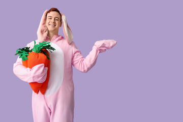 Sticker - Happy young man in Easter bunny costume with plush carrots pointing at something on lilac background