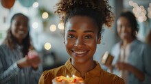 A Woman Is Smiling And Holding A Cake With Candles. She Is Surrounded By Other People, And The Atmosphere Seems To Be Celebratory