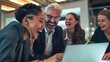 © Sasint - Happy businesspeople laughing while collaborating on a new project in an office. Group of diverse businesspeople using a laptop while working together in a modern workspace.