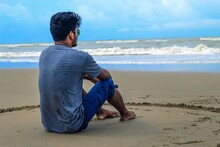 A Sad Man Sitting Down In Sand Looking At The Sea. A Young Man Sitting On A White Sand Beach During Sunrise Contemplating The Scenery And The Blue Sea Waves During His Vacation In An Idyllic Nature.