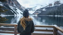 Rearview of the woman with blonde hair wearing a jacket and a winter cap, standing on the wooden deck near the lake, snowy mountains in the background, holiday or vacation, cold weather season tourism