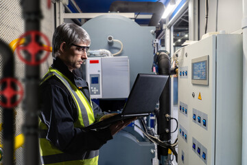Poster - Man near gas boiler equipment. Engineer sets up electric heating units. Man in engineering room of enterprise. Gray-haired engineer is holding laptop. Setting up boiler aggregate at factory