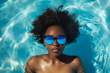 A Woman With Curly Hair Is Floating In A Pool Wearing Sunglasses. Concept Of Relaxation And Leisure, As The Woman Is Enjoying Her Time In The Water. Stylish Person Relaxing In Blue Pool