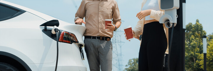 Young couple with coffee recharge EV car battery at charging station connect to electrical industrial power grid. Couple with shopping and travel using eco electric car lifestyle.Panorama Expedient