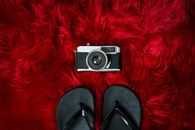 Rectangular Top View Image Of An Old Photographic Camera Next To Black Flip Flops On A Red Fur Surface