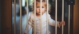 Fototapeta  - A young girl, possibly curious or confused, stands behind the sturdy bars of a jail cell. She holds onto the top of the bars and gazes directly at the camera.