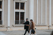 © qunica.com - Two fashionable male friends engage in a conversation while walking outside a historical building with large windows and elegant architecture.