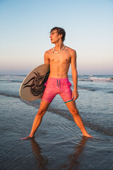 Handsome male teenage beach model standing shirtless holding his skim board while standing in the surf