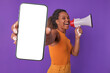 Young energetic excited African American woman promoter shows phone with white screen and shouts into megaphone calling to download social network or messenger application stands on purple background.