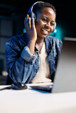 Fototapeta Tulipany - Black woman joyfully using a laptop and wireless headphones. African american female influencer multitasks, attending an online meeting while listening to music in her headset.