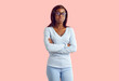 © Studio Romantic - Portrait of serious female college or high school student. Young African American woman or teenage girl wearing blue long sleeve top, jeans and glasses standing, arms folded, on pastel pink background