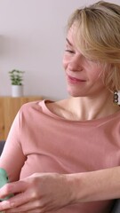 Mid adult woman relaxing on sofa using mobile phone. Happy north European female having fun watching social media content on cell phone app. Vertical HD screen video.