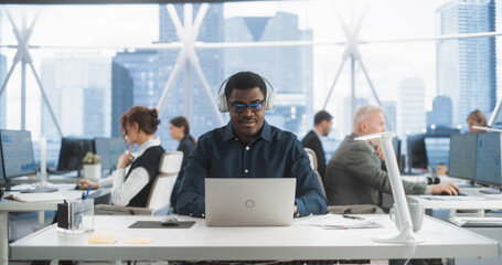 Poster - Successful Black Man Wearing Headphones And Starts Typing On Laptop Computer In Skyscraper Office In Downtown. Professional Male Software Developer Working On a New Project In Diverse Tech Startup.