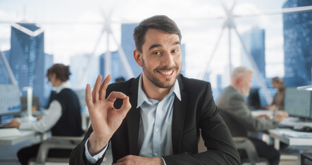 Poster - Point Of View From Camera With Handsome Entrepreneur Sitting Down On His Workplace In Diverse Skyscraper Office. Happy Caucasian Businessman Looking at Camera And Smiling, Showing OK Hand Sign.