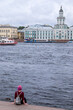 Tourist sitting on the stairs of a pier of the Neva River, with the Kunstkamera in the background, in the historic center of the city of St. Petersburg