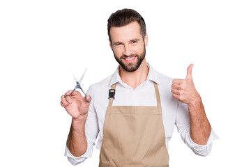 Poster - Portrait of cheerful positive barber with stubble in shirt having scissors in hand showing thumb up approve sign with finger, looking at camera, isolated on grey background