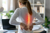 Fototapeta  - A woman working in an office has severe back pain at her workplace and touches her back. Ergonomic Alert: Woman Confronts Intense Back Pain at Desk, Urging Workplace Health Focus