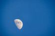 The image captures the first quarter phase of the Moon against a clear, blue sky during daytime. The Moon is detailed, with craters and maria visible, contrasting sharply with the blue expanse. The