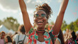 A woman with dreadlocks is smiling and holding her arms up in the air. She is wearing a colorful shirt and sunglasses. 