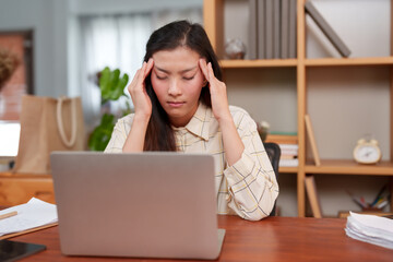 Tired employee with eyes closed, hand on temple, feeling burnout, laptop and documents present. Weary worker in moments of exhaustion, seated at desk, laptop on, hand signaling a pause.