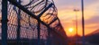 A closeup of the top edge of an iron fence with barbed wire set against a blurred background of a prison or military base during sunset Generative AI