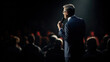 © Studio Nova - Man in a suit speaking into a microphone in front of an audience in a dark auditorium.