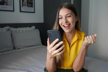 Canvas Print - Happy young Brazilian woman holding smartphone and celebrates with a clenched fist sitting on bed at home