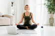 © InsideCreativeHouse - Young caucasian fit female sitting in lotus position during online training on laptop. Home yoga exercises, meditation and serenity concept