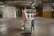 © sofiko14 - Youthful lady with her child clutching travel suitcase searching for vehicle before starting journey. Happy mom and small daughter, attired in informal wear, stroll through subterranean parking area.