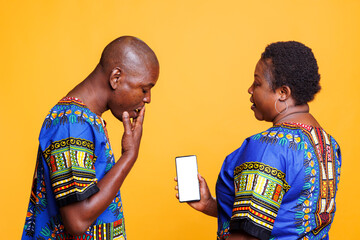 Woman showing smartphone with empty white screen with copy space to shocked man. Surprised person covering mouth with hand while looking at mobile phone with blank touchscreen mockup for app