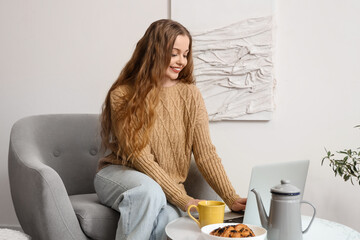 Sticker - Young woman with cup of tea in armchair using laptop at home