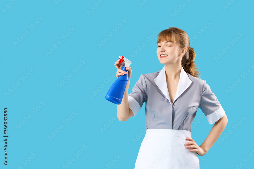 Young chambermaid with spray bottle of detergent on blue background