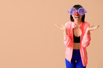 Wall Mural - Young smiling female fitness trainer with funny glasses showing her tongue on beige background. Fool's day