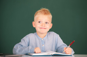 Funny kid writing in notebook in class. Child at school. Kid is learning in class on background of blackboard.