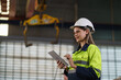 © ronnachaipark - Female Workers in green uniforms and safety helmets perform quality checks inside a metal sheet factory.