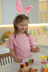 Sticker - Easter celebration. Cute girl with bunny ears painting eggs at white marble table in kitchen