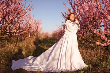 Woman blooming peach orchard. Against the backdrop of a picturesque peach orchard, a woman in a long white dress enjoys a peaceful walk in the park, surrounded by the beauty of nature.