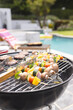 A barbecue grill is cooking skewers with vegetables and meat by a poolside with copy space