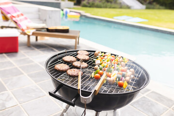 A barbecue grill is loaded with burgers and vegetable skewers, with a pool in the background