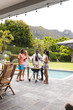 Diverse group of friends enjoy a barbecue by a pool, with mountains in the background