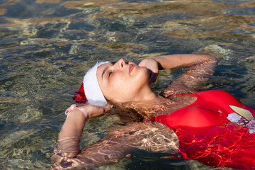 Wall Mural - Close-up portrait of a beautiful young girl with freckles, in a red swimsuit and Santa Claus hat, lying in the sea water of the Red Sea.